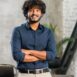 smiling-curly-middle-eastern-businessman-standing-near-desk-looking-camera-young-positive-male-student-with-modern-flat-background-proud-successful-business-owner (1)
